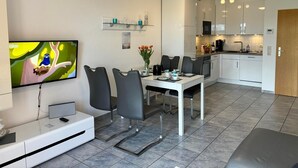 A modern kitchen with white cabinets, a dining area with a white table and black chairs, and a flat-screen TV mounted on the wall.