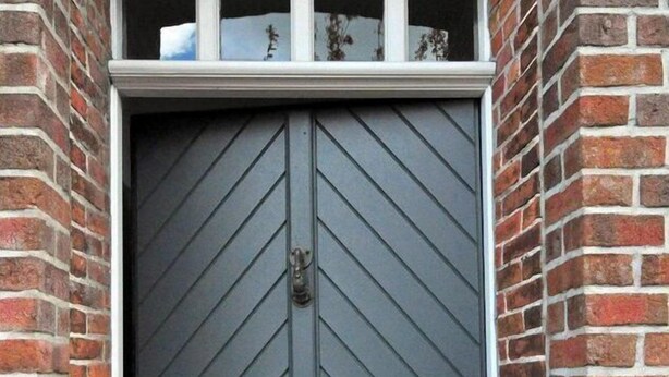 Ein dunkler, doppeltüriger Eingang mit Zickzackmuster, eingelassen in eine Backsteinmauer, oberhalb davon ein kleines Fenster.