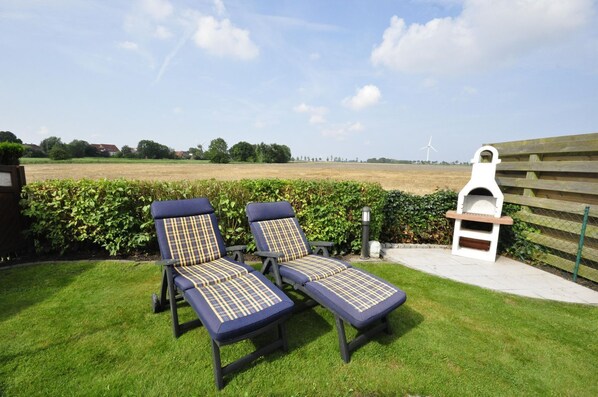 Zwei Lounge-Sessel mit kariertem Kissenmuster, ein Grill und ein Blick auf ein Feld sowie Windkraftanlagen im Hintergrund.