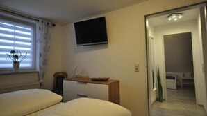 Une chambre d’hôtel avec un lit, un téléviseur fixé au mur, une petite table d’appoint en bois avec un bol, une plante en pot et une fenêtre avec des stores.