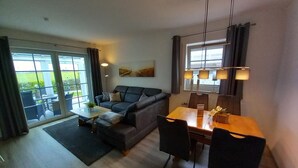 Un salon moderne avec un canapé, une table à manger et des chaises. On y trouve une grande fenêtre avec des rideaux, une petite table sur laquelle est posée une plante, et une lampe fixée au mur.
