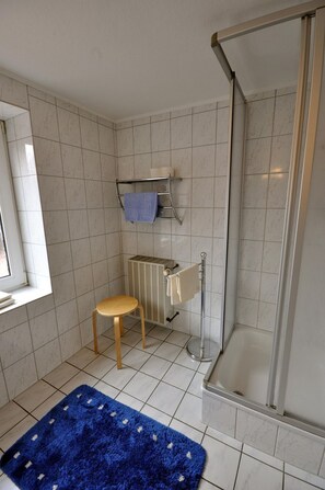 Uma casa de banho com paredes revestidas a mosaico branco, chuveiro, sanita e lavatório com porta-toalhas, e um tapete azul no chão.