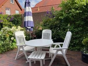 Une terrasse avec une table ronde et quatre chaises blanches, entourée d’une végétation luxuriante et d’un mur de briques.
