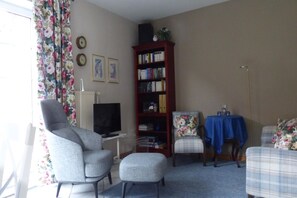 Un salon avec un fauteuil gris, une petite table recouverte d’une nappe bleue, une bibliothèque et un téléviseur.