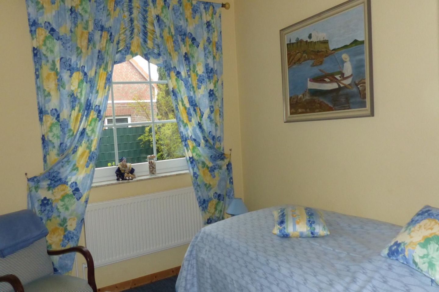 A room with a bed, a window with floral curtains, a radiator, and a painting of a boat on the wall.