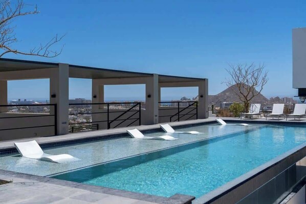 A rooftop infinity pool with white lounge chairs, a clear view of the cityscape, and a mountain in the background.