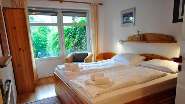 A neatly made bed with white linens and towels, a wooden headboard, a bedside table with a lamp, and a window with curtains.