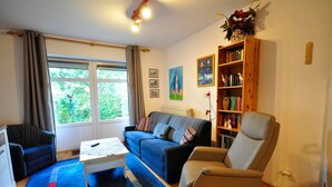 A living room with a sofa, armchair, coffee table, bookshelf, and a window with curtains.