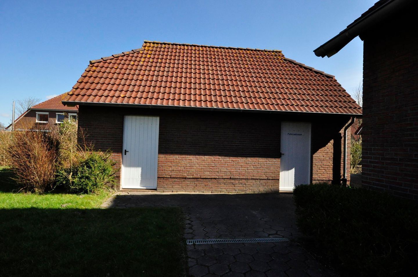 A two-story brick building with a red-tiled roof, two white doors, and a small garden area with grass and shrubs.