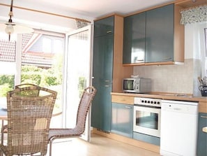 Cocina equipada con microondas, horno y lavavajillas. Hay una silla de mimbre y una mesa.