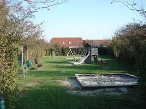 Un parque infantil con tobogán y columpios, rodeado de árboles y un edificio al fondo.