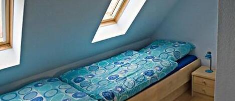 A small, single-bed room with a sloped ceiling and skylight, wooden flooring, and a bedside table with a lamp.
