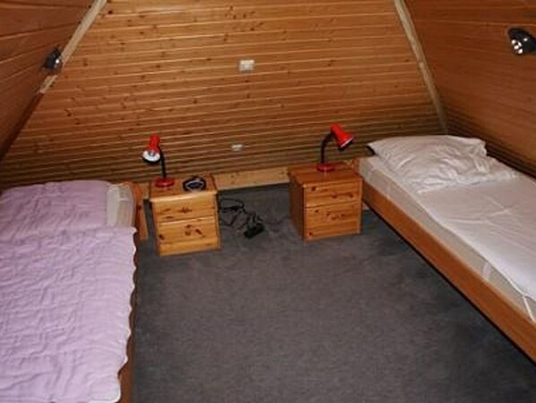 A small, cozy room with a sloped ceiling, two wooden drawers with red lamps, a single bed with a white mattress, and a purple blanket.