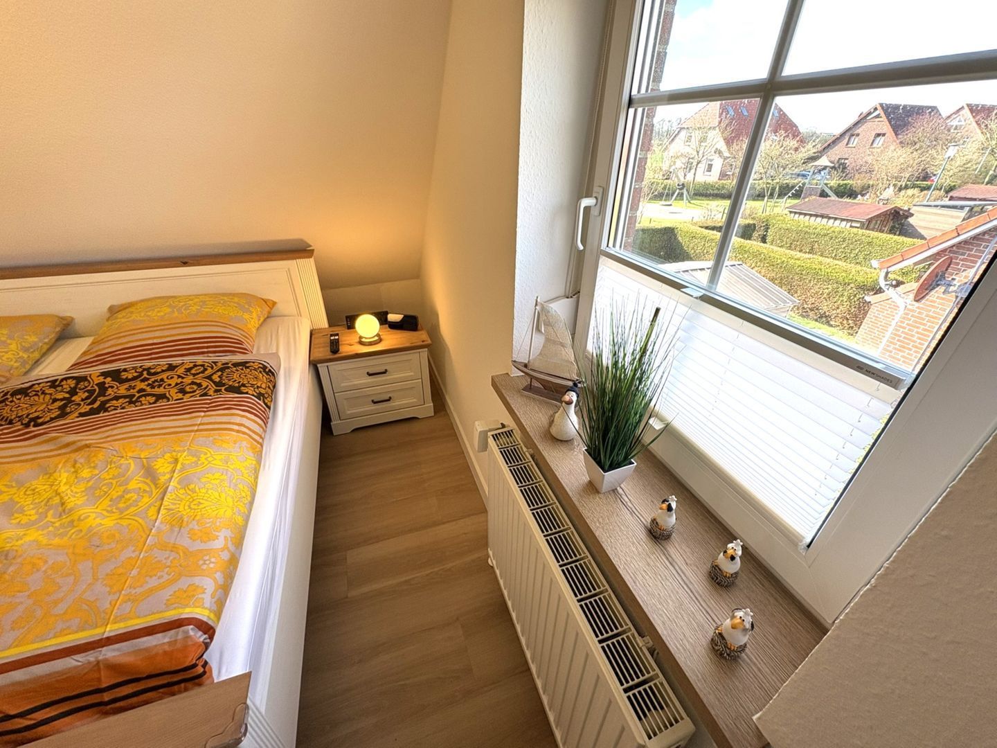 A bedroom with a bed, bedside table, lamp, radiator, window, and view of the outside.