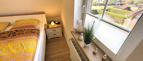 Un dormitorio con cama, mesita de noche, lámpara, radiador, ventana y vistas al exterior.