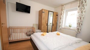 Ein Schlafzimmer mit einem Babybett, einem Bett, einem Kleiderschrank, einem Fernseher und einem Fenster mit Vorhängen.