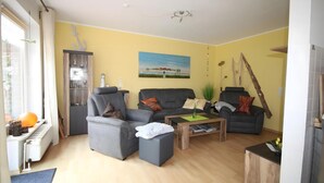 Ein Wohnzimmer mit Sofa, Sessel und Couchtisch. Ein Gemälde hängt an der Wand, und ein Fenster ist mit Vorhängen versehen.