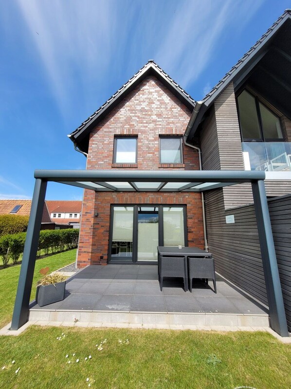 Een huis met twee verdiepingen, een bakstenen gevel, grote ramen en een overdekte patio met buitenmeubels.