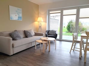 A living room with a sofa, a chair, a small table, and a lamp. There is a large window with blinds.