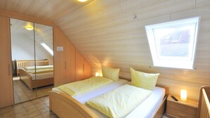 A bedroom with a wooden ceiling, a bed with yellow bedding, a nightstand, a mirror, and a window with a view of a building. - Vacation home Uferstraße 19b (Wittmund)