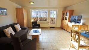 A living room with a sofa, coffee table, and TV in a well-lit space with wooden flooring and large windows. - Vacation home Uferstraße 19b (Wittmund)