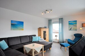 A modern living room with a black sofa, a small wooden table, and a blue armchair. There are two paintings on the walls, a wooden cabinet, and a window with curtains.