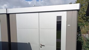 Un abri en métal gris, doté d’une porte et d’une fenêtre, est installé sur une surface pavée, avec de l’herbe et une clôture en arrière-plan.