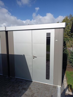 A grey metal shed with a door and a window, set on a paved surface with grass and a fence in the background. - Vacation w. Int Seeburger Winkel 5 OG - Holiday apartment Int Seeburger Winkel 5 OG (Wittmund)