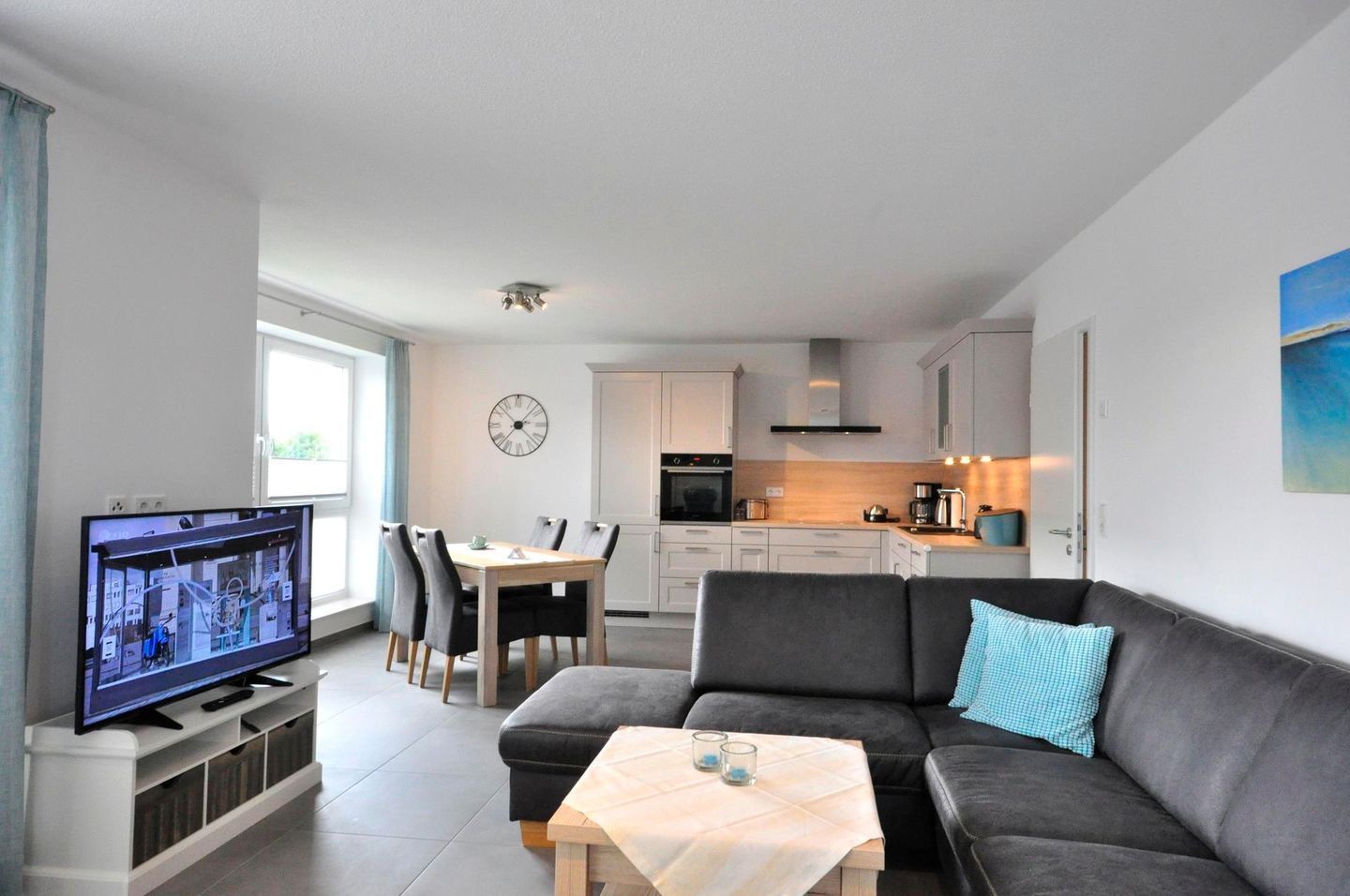 A modern living room with a flat-screen TV, a grey sofa, a wooden coffee table, and a dining area with a wooden table and chairs.