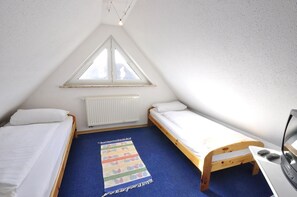 Ein kleiner, dreieckiger Raum mit zwei Einzelbetten, einem Fenster, einer Heizung und einem Schreibtisch mit einem Computer.