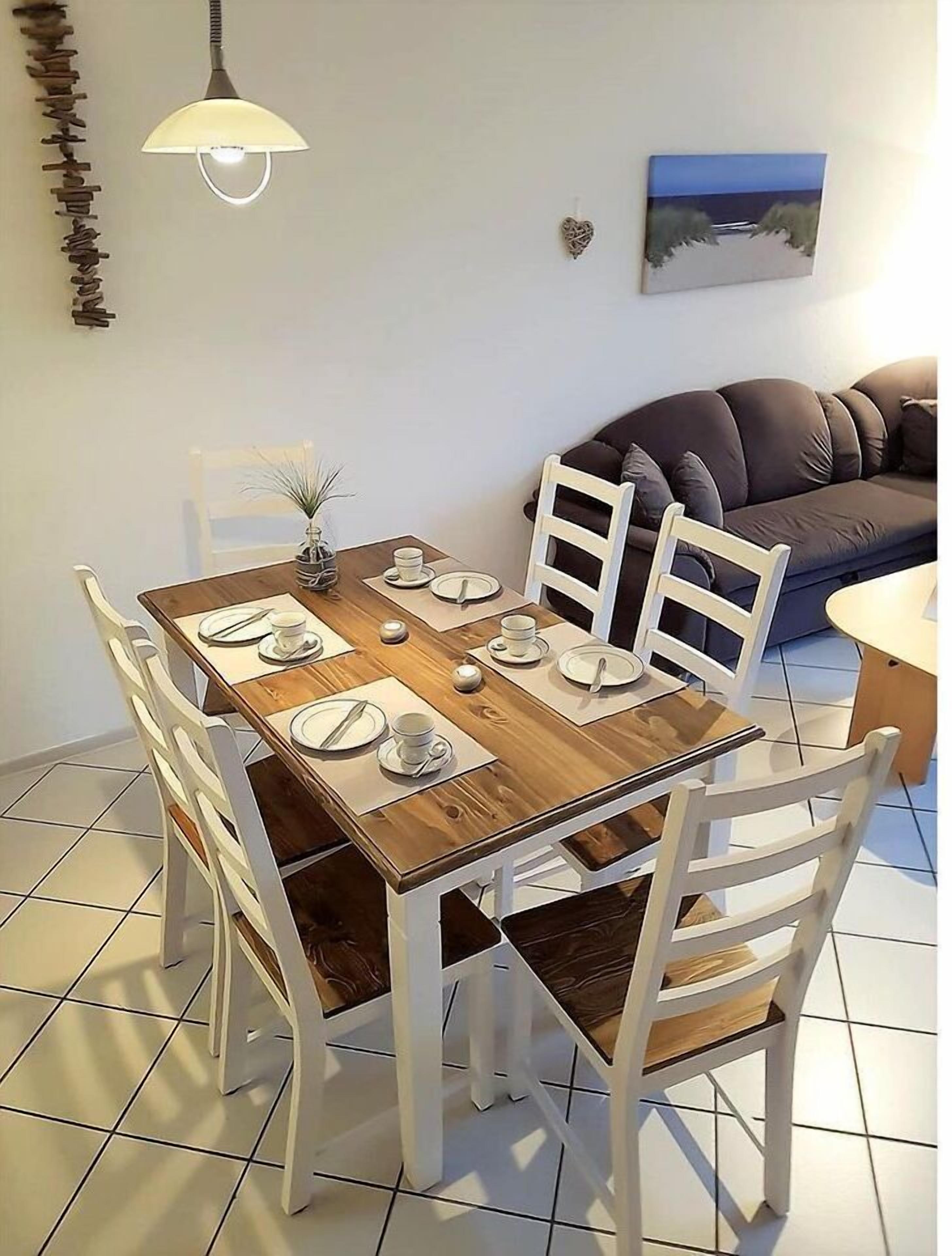 Een eetruimte met een houten tafel gedekt voor vier personen, witte stoelen, een hanglamp en een schilderij aan de muur.
