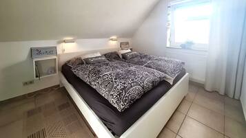 Ein Schlafzimmer mit einem Bett, zwei Nachttischen, einem an der Wand montierten Regal, einem Fenster mit Vorhängen und einer Heizung.