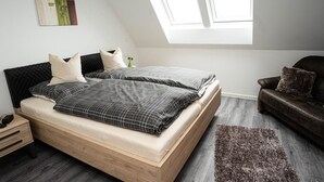 A bedroom with a bed, a nightstand, a wooden dresser, a brown leather sofa, a window, and a painting on the wall. - Vacation home Krabbenpadd 9 "Find's Huus" - Holiday home "Find's Huus" Krabbenpadd 9 (Wittmund)