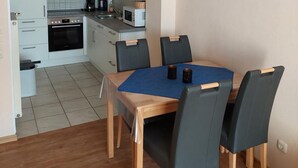 A compact kitchen with white cabinets, a microwave, and a stove. There is a dining area with a wooden table and four chairs. - Vacation home Mühlenblick 41 No. 3 (Wittmund)