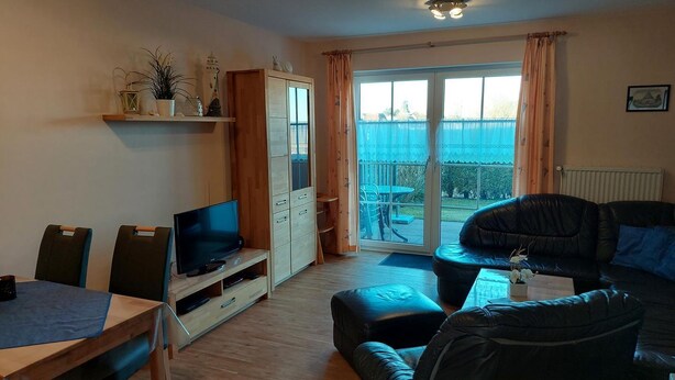 A living room with a TV, a sofa, a dining table with chairs, a cabinet, and a sliding glass door leading to an outdoor area. - Vacation home Mühlenblick 41 No. 3 (Wittmund)