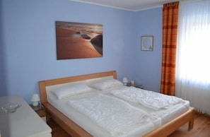 Een slaapkamer met een houten bed, wit beddengoed, een nachtkastje, een lamp en een schilderij van een strandgezicht.