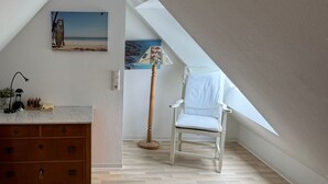 Une pièce avec un plafond en pente, une chaise blanche, une commode en bois et un tableau représentant une scène de plage.