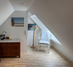 Une pièce avec un plafond en pente, une chaise blanche, une commode en bois et un tableau représentant une scène de plage.