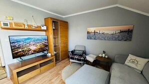 Un salon doté d’un téléviseur à écran plat, d’un meuble de rangement en bois, d’un canapé gris, d’un fauteuil à motifs et d’un tableau représentant une scène de plage.