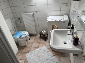 Et badeværelse med toilet, radiator, vask og et lille bord med toiletpapir.