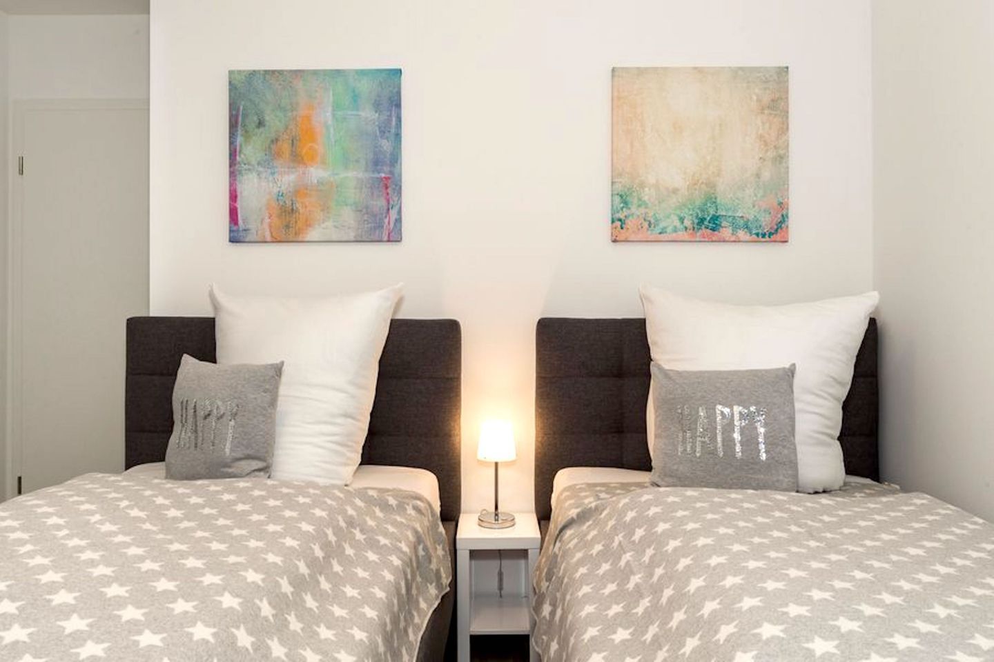 Une chambre double avec deux lits, chacun recouvert d’une couvre-lit grise à motifs étoilés, agrémentée de coussins blancs et d’une petite table de chevet avec une lampe.