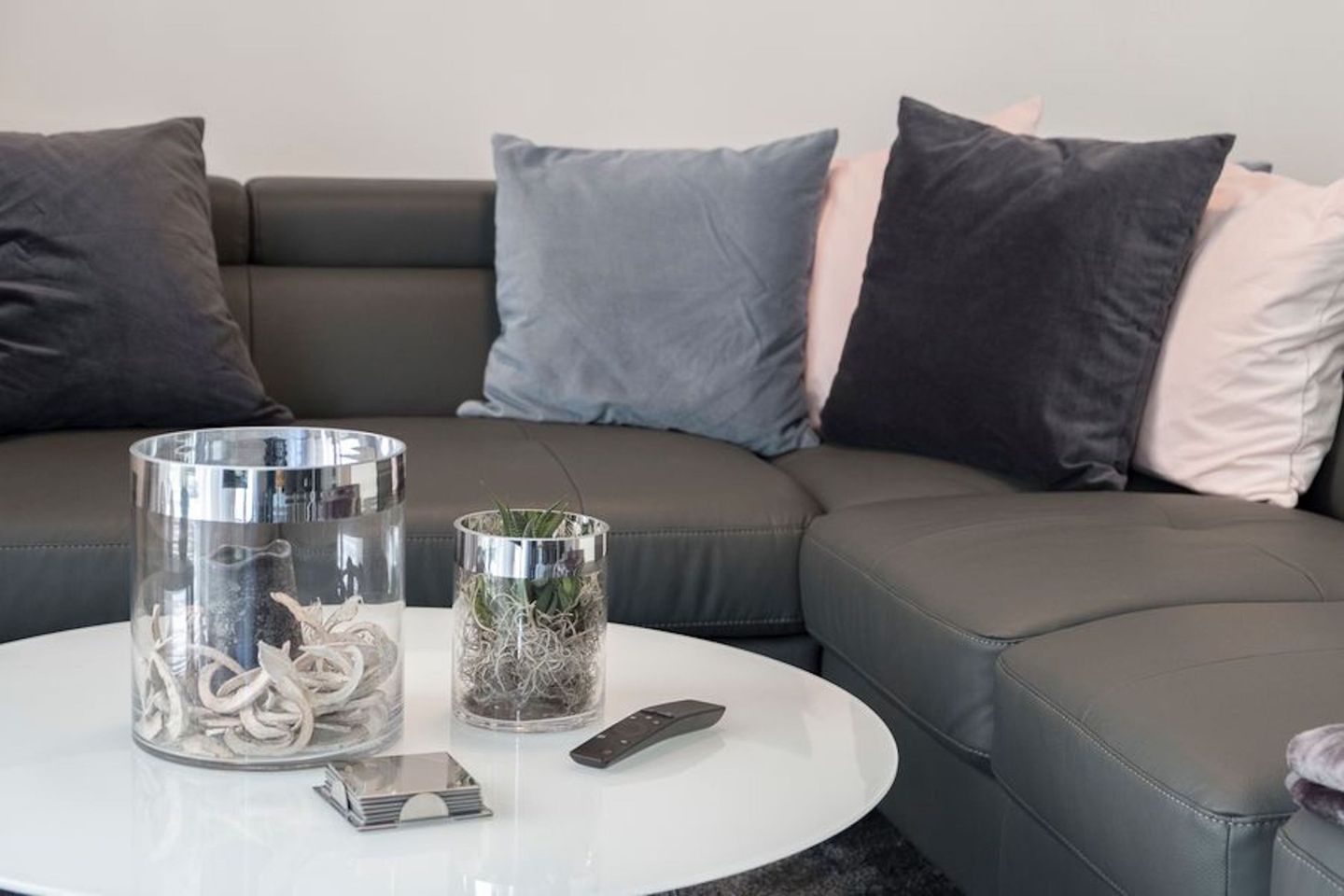 Ein modernes Wohnzimmer mit einem grauen Ledersofa, zwei Glasvasen mit Dekoration, einer Fernbedienung und einem kleinen Tablett auf einem weißen Couchtisch.