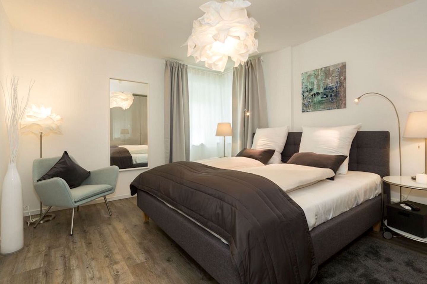 A modern hotel room with a large bed, a sitting area with a chair, a bedside table, and a large window with curtains.
