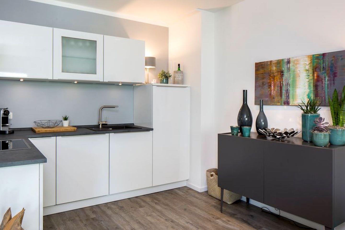 Una cocina moderna con armarios blancos, encimera negra y suelo de madera. Hay un lavamanos con grifo, una cafetera y una pequeña planta en la encimera.