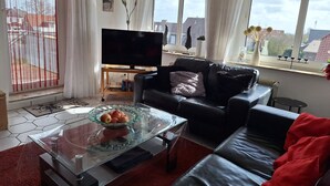 Ein Wohnzimmer mit einer Ledersofa, einem Glastisch mit Früchten, einem Fernseher auf einem Holzboden und großen Fenstern mit Vorhängen.