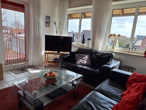 A living room with a leather sofa, a glass coffee table with fruit, a TV on a wooden stand, and large windows with curtains.