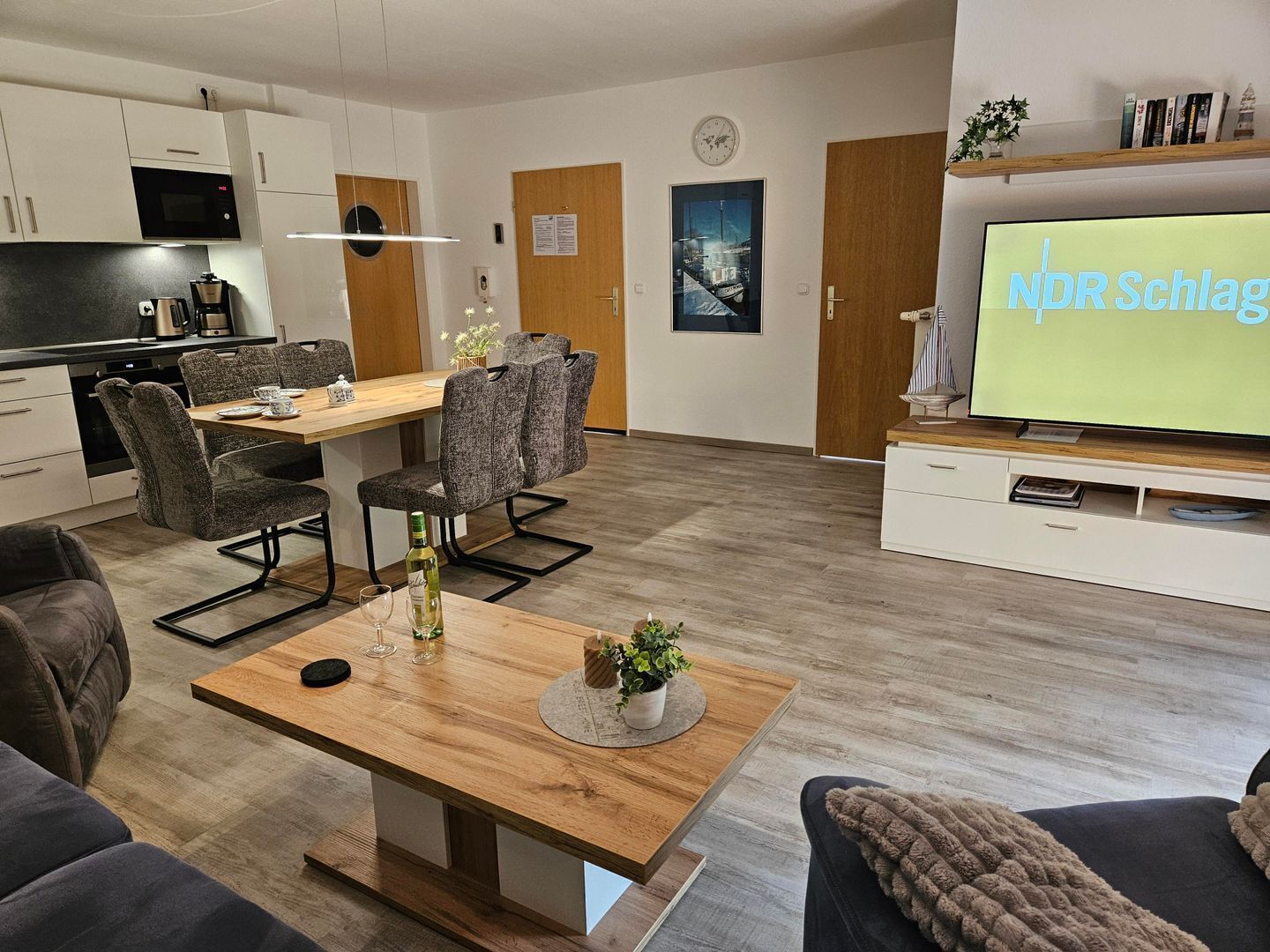 Ein modernes Wohnzimmer mit Couchtisch aus Holz, grau gepolsterten Sesseln, einem Flachbildfernseher und einer Küchenzeile mit weißen Schränken und einer Mikrowelle.