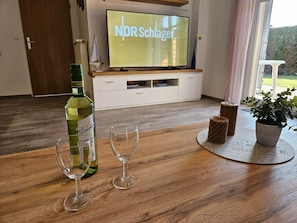 Un salon avec une table en bois, une bouteille de vin Blanquette, deux verres, une plante en pot et un téléviseur diffusant l’émission « NDR Schlager ».