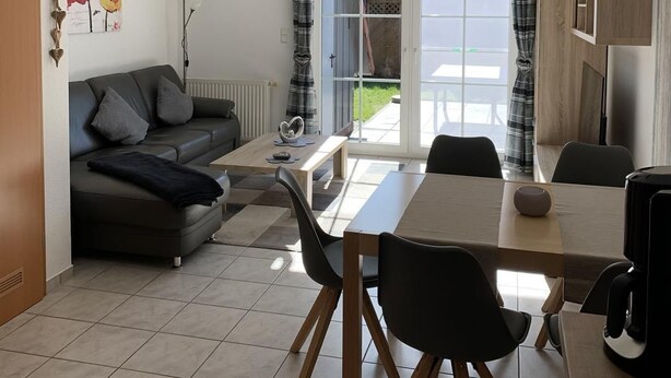 A modern living room with a sofa, coffee table, and dining table. There is a large window with curtains, a ceiling light, and a door leading outside. - Vacation cottage Mittelweg 41a (Wittmund)