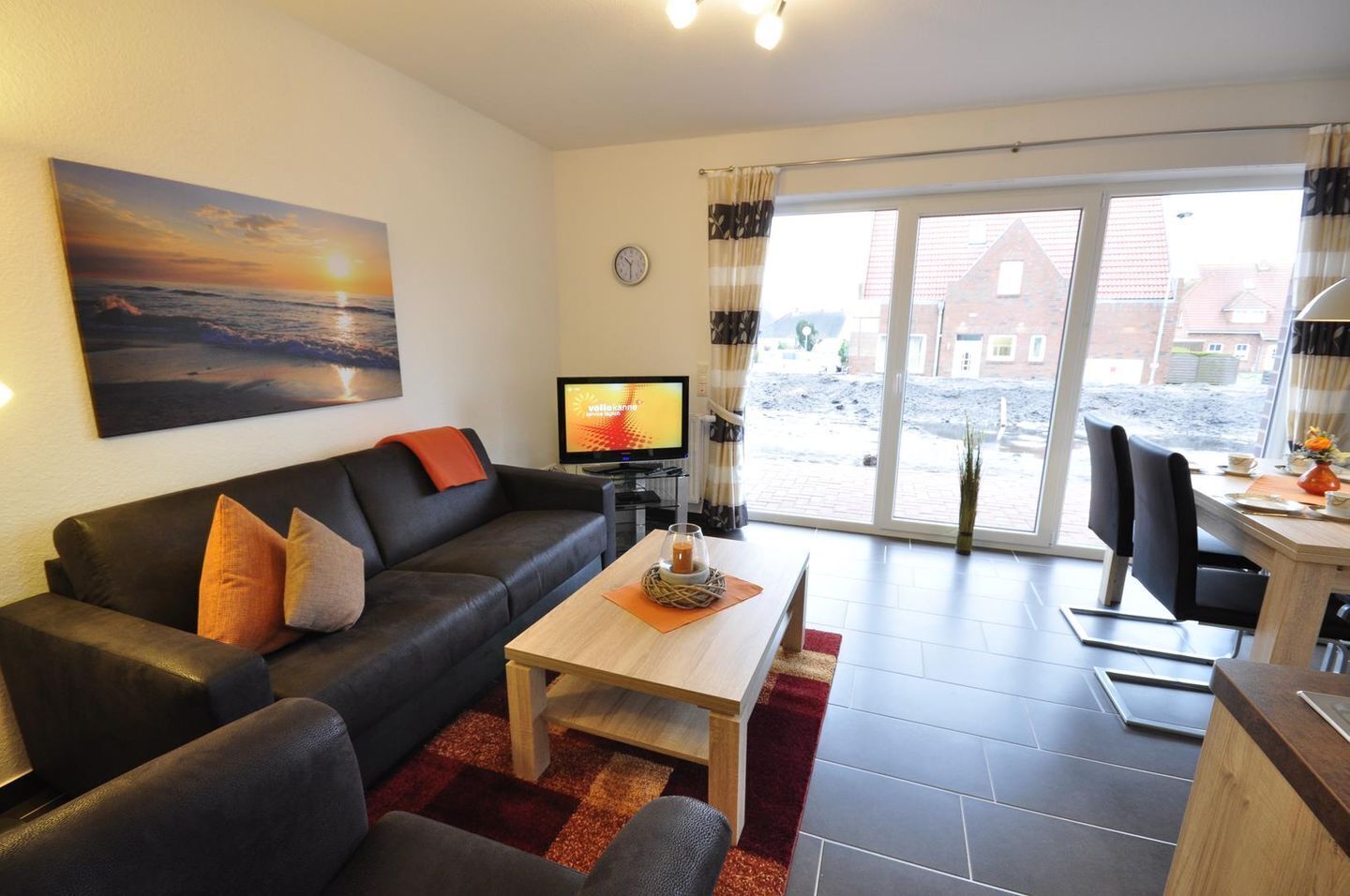 Een moderne woonkamer met een bank, salontafel en een televisie. Er is een eethoek met stoelen en een tafel. De kamer heeft grote ramen met uitzicht op het buitenland.